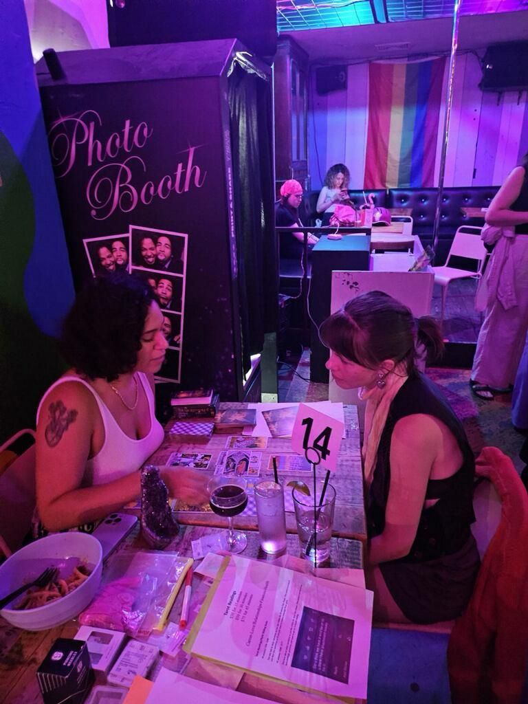 Kaide giving a tarot reading at a bar event, with a photo booth and a Pride flag in the background