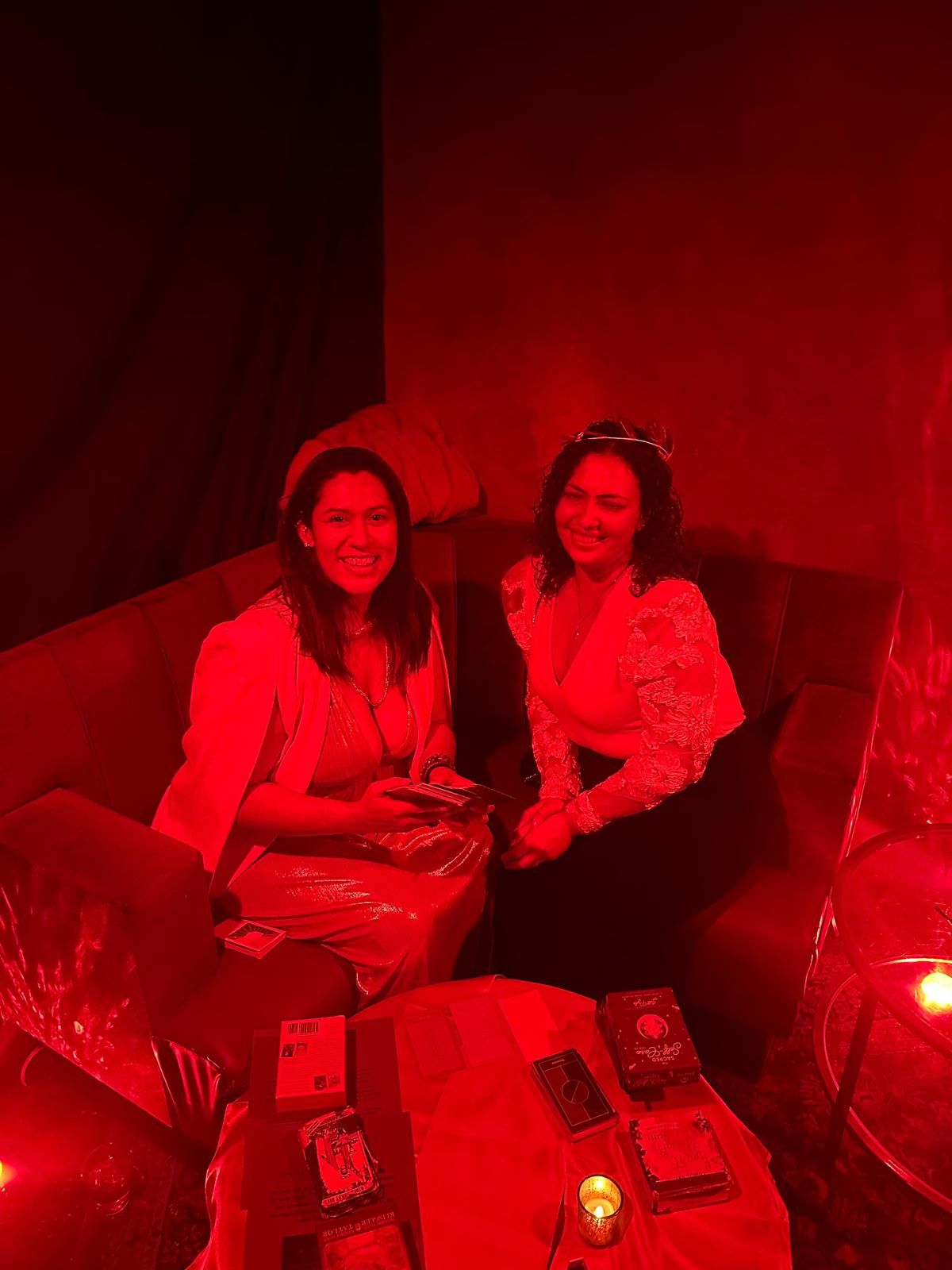 Kaide and a guest smiling on a sofa in red light, with tarot decks and a candle on the table in front of them